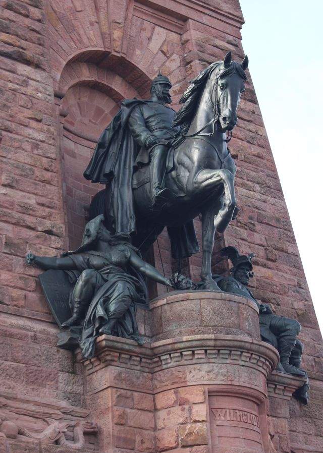 Kaiser Wilhelm I auf einem Pferd. Die Skulptur zeigt ihn mit Pickelhaube. Flankiert wird er von zwei allegorischen Figuren.