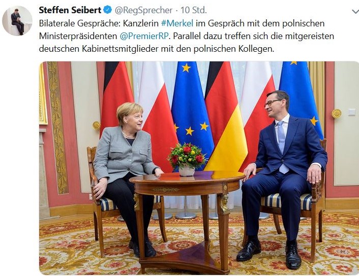 Angela Merkel mit gruem Blazer und Mateusz Morawiecki im dunkleblauen Anzug an einem runden Holztisch. Auf diesem stehen Blumen, im Hintergrund die polnische und deutsche sowie die EU-Fahne.