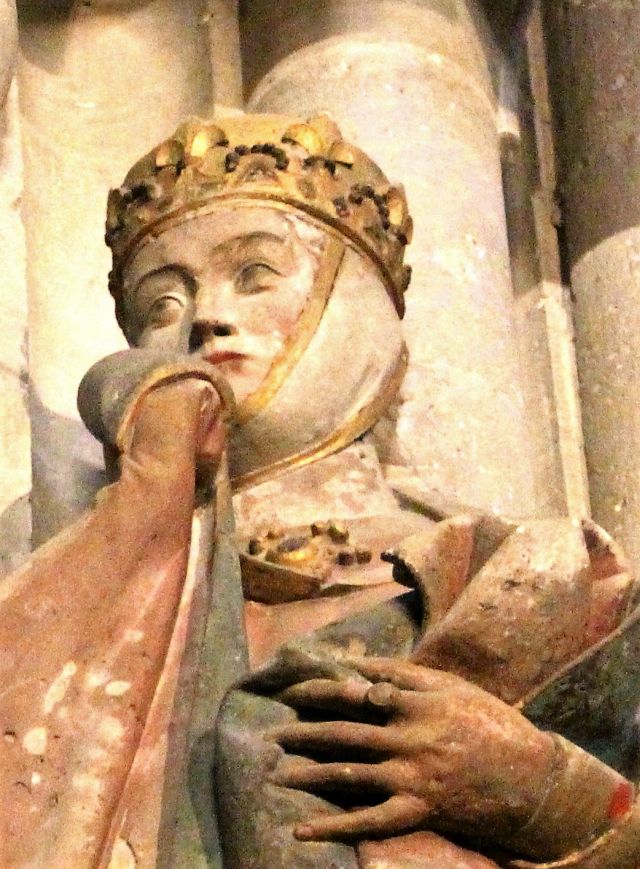 Der Kopf einer Frau mit einer goldfarbenen Krone. Sie zieht den rechten Teil des Mantelkragens ins Gesicht.
