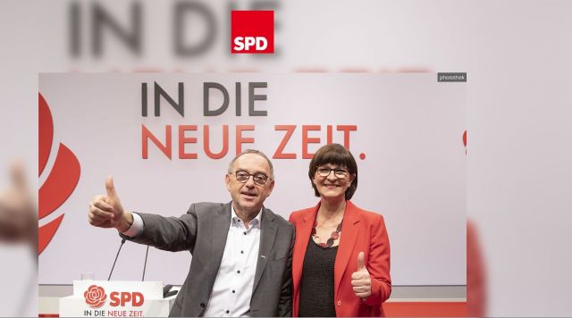 Norbert Walter-Borjans mit hellgrauem Jacket und offenem weißen Hemd. Saskia Esken mit roter Jacke. Beide zeigen die Daumen nach oben. Dahinter der Schriftzug "In die neue Zeit".
