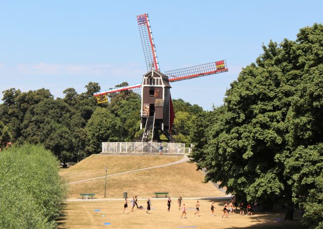 Eine Windmühle aus Holz auf einem kleinen Hügel. Im Vordergrund Menschen in einem Park. 