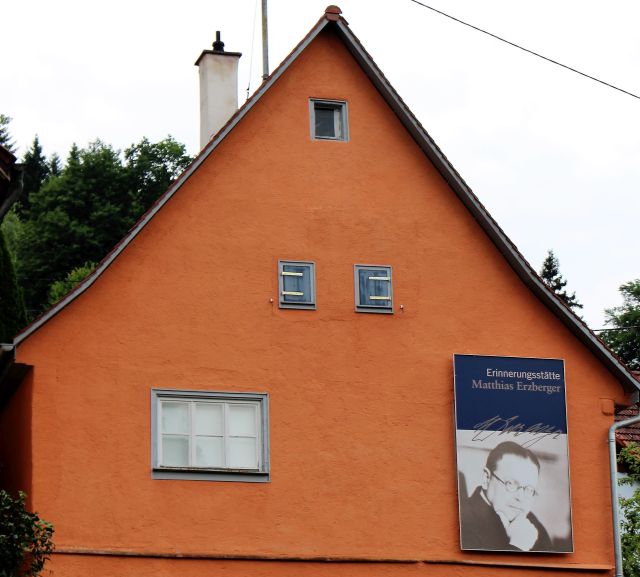 Geburtshaus Erzbergers. Fassade in einem rötlichen Ton. Kleine Fenster. Ein Plakat weist mit einer Porträtaufnahme Erzbergers und dem Hinweis auf die Gedenkstätte.