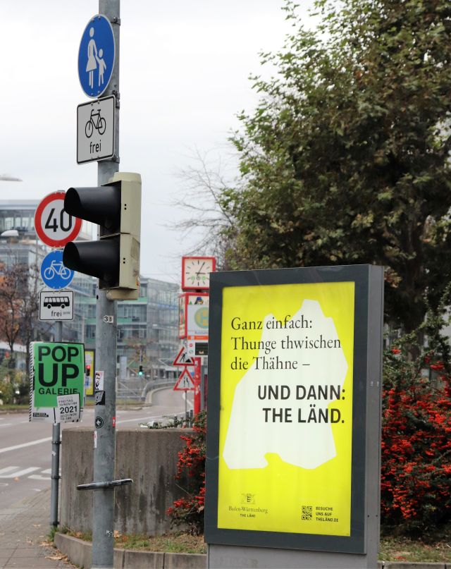 Gelbes Werbeplakat mit schwarzer Schrift "Ganz einfach Thunge zwischen die Thähne – und dann The Länd“. Im Umfeld eine Unzahl von Verkehrs- und anderen Hinweisschildern.