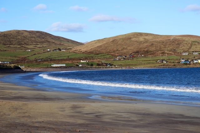 Blaues Meer mit auflaufender Welle. Der weite Sandstrand lässt erkennen, dass Ebbe herrscht. Im Hintergrund an der Küste einzelne Gebäude.
