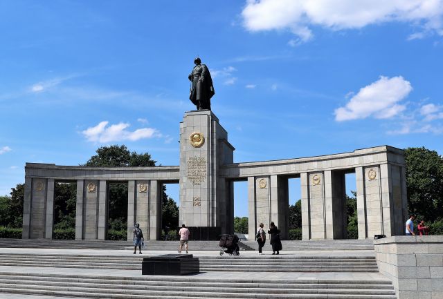 Blick auf die Frontseite des Sowjetischen Ehrenmals. Mehrere Besucher vor dem Denkmal. In der Mitte eine schwarze Metallskulptur, die einen Soldaten mit Mantel, Helm und geschultertem Gewehr zeigt. Links und rechts eine bogenförmige Steinstruktur mit Durchgängen.
