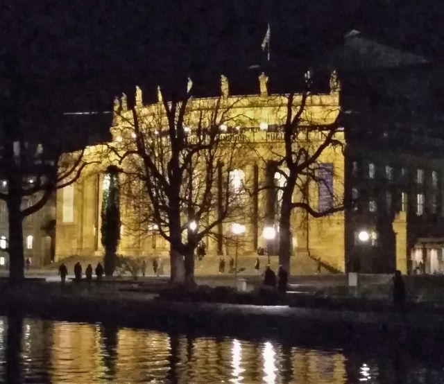 Gebäude der Staatsoper in Stuttgart bei Nacht. Einzelne Lampen schaffen etwas Helligkeit. Davor funkeln sie im Eckensee.