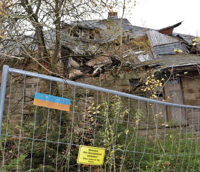 Ein Bauzaun mit dem gelben Schild "Betreten des Grundstücks verboten! Eltern haften für ihre Kinder!" Im Gebäude, das sich dahinter erhebt, ist der Dachstuhl eingestürzt.