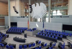 Das leere Plenum des Deutschen Bundestags im Reichtag. Der Bundesadler an der Rückseite, davor die deutsche und die EU-Flagge. Blaue Sitze im großen Rund.