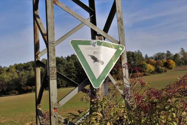 Ein verbogenes und verblichenes Schild Naturschutzgebiet hängt unmittelbar vor einem Strommasten. In größerer Entfernung ein Waldstück.