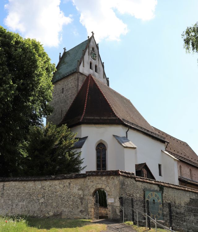 Ein weißes Kirchengebäude mit einem rötlichen Ziegeldach. Der viereckige Turm wurde mit Natursteinquadern gemauert. Das Dach des Turms ist dunkel. Eine Uhr mit goldenen Lettern zeigt 17 Uhr 10. 