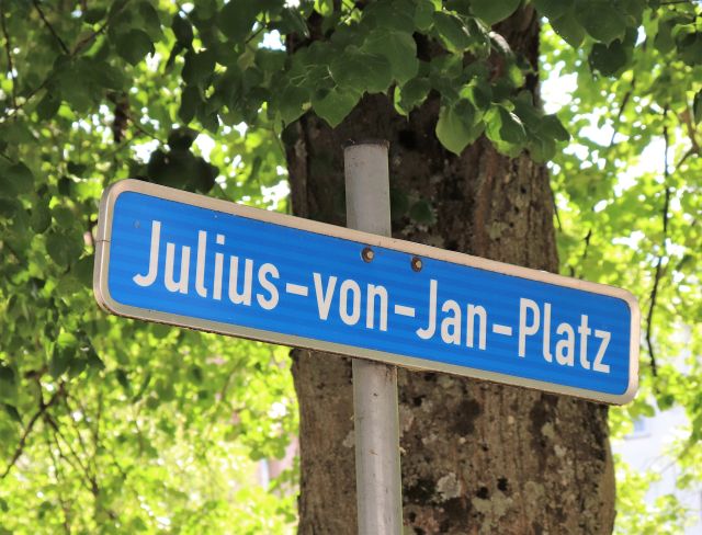 An einem silbernfarbenen Pfahl ist ein blaues Schild befestigt. Text: Julius-von-Jan-Platz.