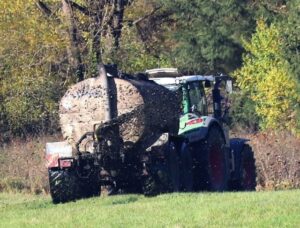 Gezogen von einem grünen Traktor wird aus dem Tankanhänger gut erkennbar Gülle auf das Grünland geschleudert.
