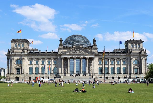 Vorderansicht des Reichstags mit deutschen Flaggen und EU-Fahne. Zahlreiche Menschen sitzen auf der Grünfläche bzw. gehen darüber. Der Himmel ist blau mit wenigen weißen Wolken.