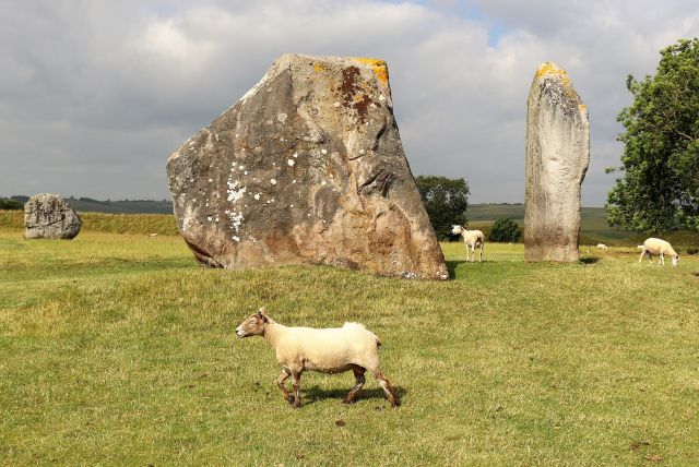 Zwei gewaltige Steine bzw. Felsbrocken stehen aufrecht auf einer grünen Wiese. Dazwischen Schafe mit heller Wolle.
