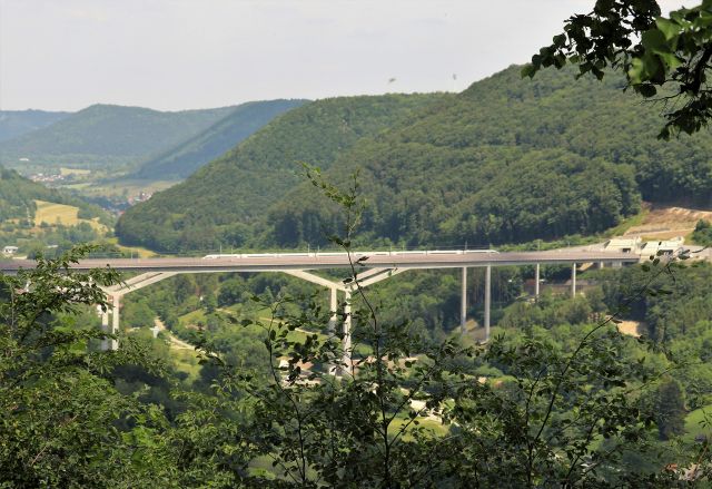 Eine moderne Eisenbahnbrücke aus hellem Beton, über die gerade ein silberner ICE fährt. Rechts ist über einem bewaldeten Hang die nächste Tunneleinfahrt zu sehen.