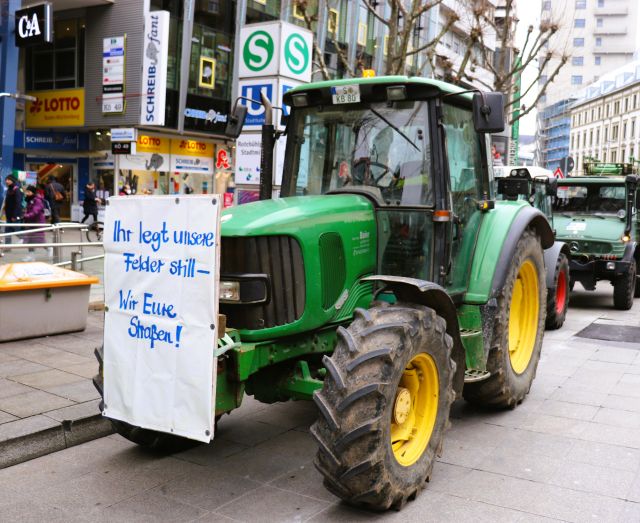 Ein grüner Traktoor mit einem weißen Plakat: "„Ihr legt unsere Felder still – Wir Eure Straßen!“