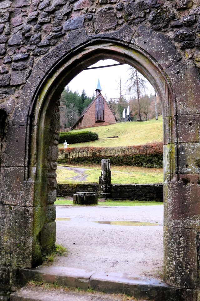 Blick durch eine Öffnung auf eine Kapelle, die oberhalb der Klosterruine am Hang steht. Sie hat einen Dachreiter mit einem Kreuz.