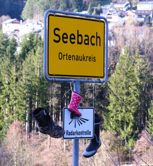 Gelbes Ortsschild mit der Aufschrift 'Seebach. Ortenaukreis'. Darunter ein kleines weißes Schild 'Radarkontrolle'. Am Ortschild hängen verschiedenfarbige Gummistiefel.