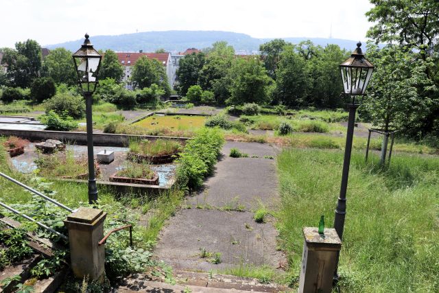 Verwahrlostes Außengelände. Ungepflegte Grasflächen und Teile eines früheren Kaskadenbrunnens. Vorne zwei Lampen im historischen Stil.