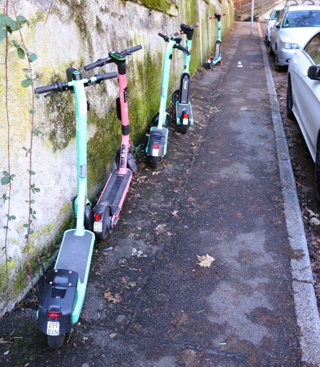 Fünf grüne und rote E-Scooter stehen an der Wand, die einen Gehweg gegen höheres Gelände absichert.