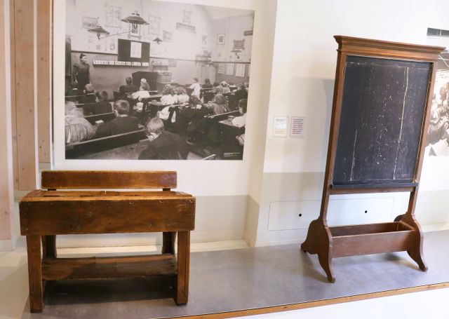 Eine hölzerne Schulbank mit Tisch, eine große Schiefertafel und das Foto einer Schulklasse.
