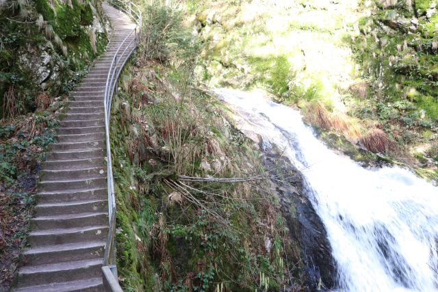 Links führen steinerne Stufen mit einem Geländer nach oben, parallel zum Wasserfall.