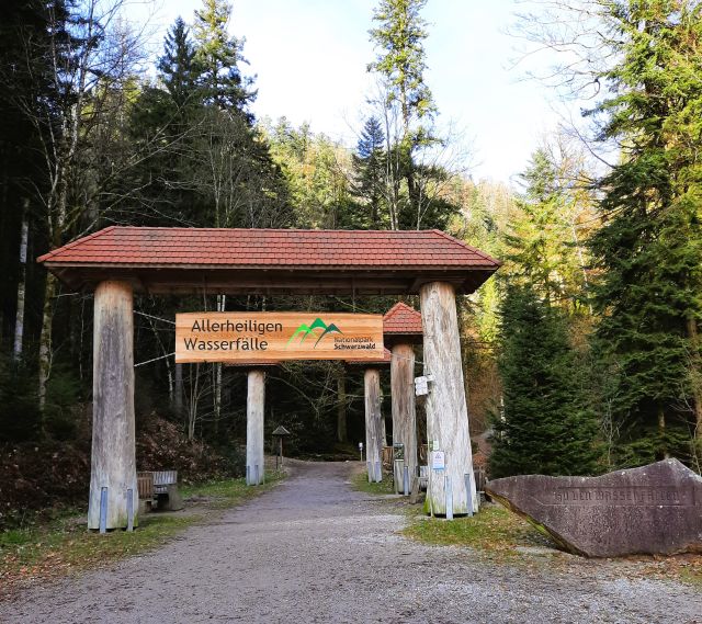 Ein Tor aus Baumstämmen mit einem kleinen Ziegeldach und einem hölzernen Schild mit der Aufschrift "Allerheiligen Wasserfälle. Nationalpark Schwarzwald".