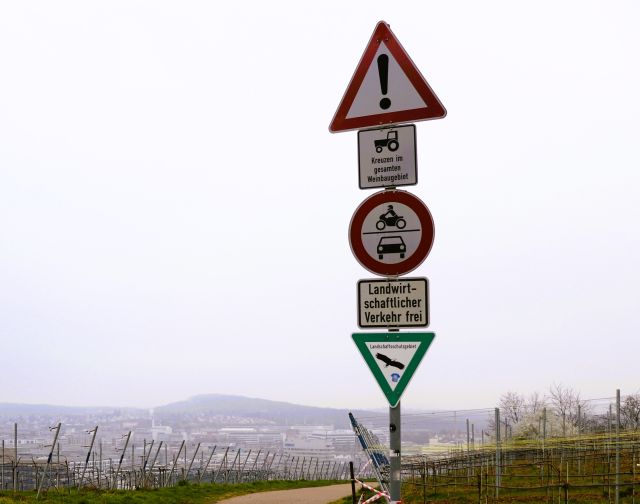 An einem Metallpfahl sind mehrere Schilder befestigt, so Landschaftsschutzgebiet, Verbotsschild für Fahrzeuge, frei für Traktoren. Dahinter einige Weinstöcke und Industrieansiedlungen.