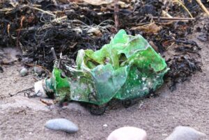Eine grüne recht zernitterte Plastikflasche mit Löchern liegt am Sansstrand zwischen einigen rundlichen Steinen und angeschwemmtem Seetang.