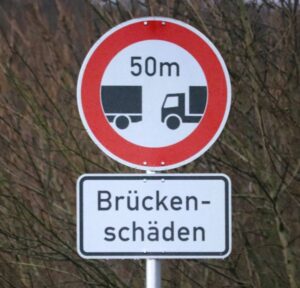 Im Bild zu sehen: Ein Schild mit dem Text 'Brückenschäden' in schwarzen Buchstaben auf weißem Grund. Darüber ein rundes Schild mit rotem Rand und zwei Lkw mit dem Abstandsgebot von 50 Metern.